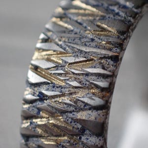 May include: Close-up of a silver ring with a textured surface. The surface is patterned with a series of raised lines that create a geometric design. The lines are a light gold colour, and the background is a dark silver colour.