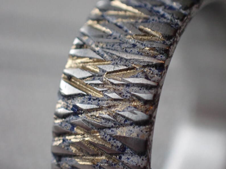 May include: Close-up of a silver ring with a textured surface. The surface is patterned with a series of raised lines that create a geometric design. The lines are a light gold colour, and the background is a dark silver colour.