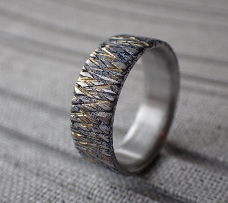 May include: A silver and gold wedding band with a textured, geometric pattern. The band is made of two metals, with the gold accents creating a contrasting design.