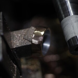 May include: Close-up of a textured, gold-coloured ring with a faceted design. The ring is set against a blurred background of metalworking tools, highlighting the craftsmanship of the jewellery.