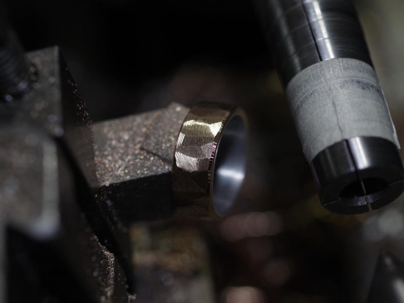 May include: Close-up of a textured, gold-coloured ring with a faceted design. The ring is set against a blurred background of metalworking tools, highlighting the craftsmanship of the jewellery.