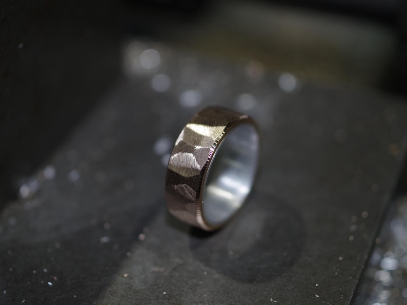 May include: A hammered metal ring with a textured surface. The ring has a silver interior and a bronze exterior. The ring is set against a dark, textured background, highlighting its metallic sheen.