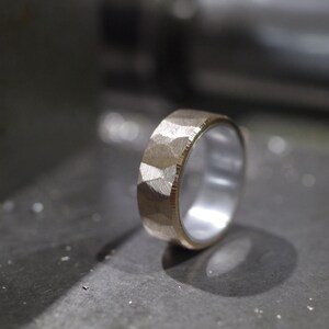 May include: A textured gold ring with a silver interior, shown in close-up. The ring's surface has a faceted design. The ring is set against a blurred, industrial background.