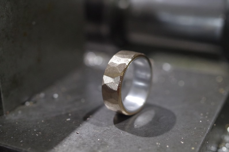 May include: A textured gold ring with a silver interior, shown in close-up. The ring's surface has a faceted design. The ring is set against a blurred, industrial background.