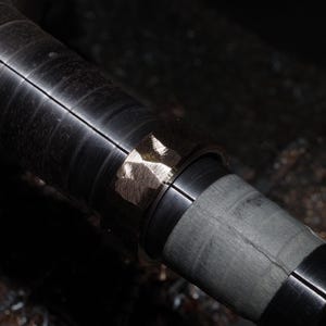 May include: Close-up of a metal lathe component. The cylindrical piece is dark grey with a series of grooves. A section of the metal has a lighter, textured appearance, possibly from machining or finishing.