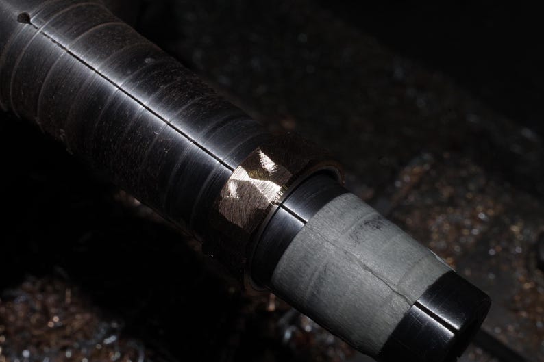 May include: Close-up of a metal lathe component. The cylindrical piece is dark grey with a series of grooves. A section of the metal has a lighter, textured appearance, possibly from machining or finishing.