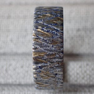 May include: A close-up of a silver ring with a textured pattern of blue and gold lines. The ring is made of metal and has a polished finish.