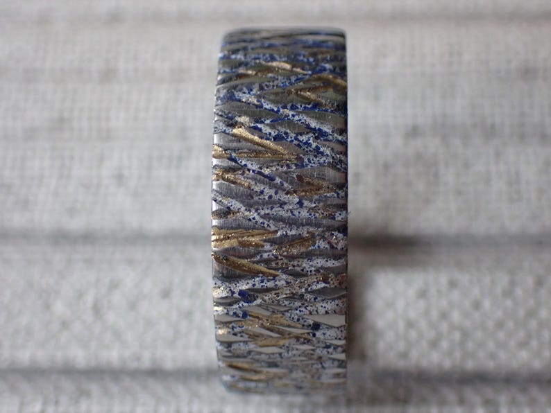 May include: A close-up of a silver ring with a textured pattern of blue and gold lines. The ring is made of metal and has a polished finish.