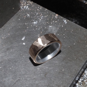 May include: A textured, rose gold ring with a silver interior. The ring is set on a dark grey surface, with small metal shavings scattered around it. The ring's surface has a geometric pattern.