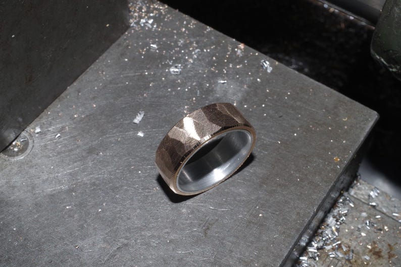 May include: A textured, rose gold ring with a silver interior. The ring is set on a dark grey surface, with small metal shavings scattered around it. The ring's surface has a geometric pattern.