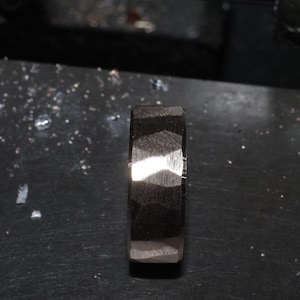 May include: A dark, textured ring with a geometric pattern. The ring is made of a dark material, possibly metal, and has a faceted surface that catches the light. The ring is displayed on a dark surface.
