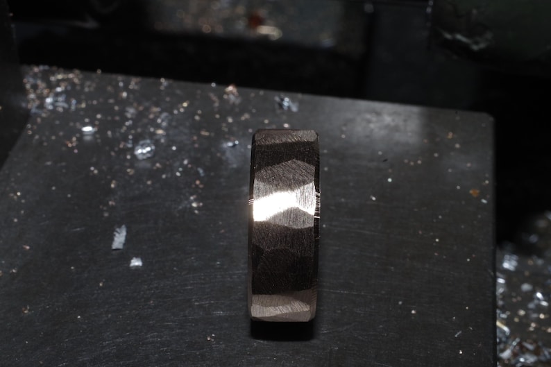May include: A dark, textured ring with a geometric pattern. The ring is made of a dark material, possibly metal, and has a faceted surface that catches the light. The ring is displayed on a dark surface.