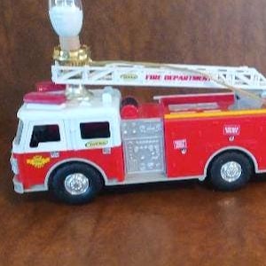May include: A red and white toy fire truck with a ladder and a light on top. The truck has the words "Fire Department" on the side. The truck is plugged in and ready to play.