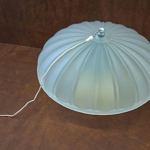 May include: A frosted glass pendant light fixture with a ribbed, dome-shaped shade. The light is a soft, pale blue-green color and has a white cord and a metal chain. The fixture is on a brown surface.
