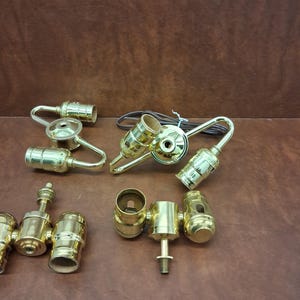 May include: A collection of brass lamp parts, including sockets, hooks, and connectors, arranged on a brown surface. The components vary in shape and size, with a shiny, metallic finish. A brown electrical cord is also present.