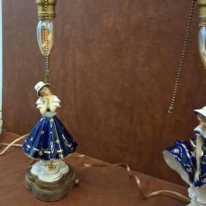 May include: Two vintage porcelain lamps featuring a woman figurine with a blue dress and white hat. The lamps have brass fixtures and a decorative base. The lamps are on a brown surface.