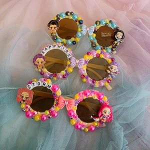 Puede incluir: Una colección de gafas de sol decorativas con lentes marrones. Cada par presenta un diseño único con cuentas de colores, lazos y pequeñas figuras de personajes. Las gafas de sol deletrean "SAPPHIRE" y "STORM".