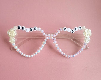 Może przedstawiać: Białe plastikowe okulary z tęczowymi koralikami i białymi kwiatami. Okulary mają napis "BRIDE VIBES" czarnymi literami.