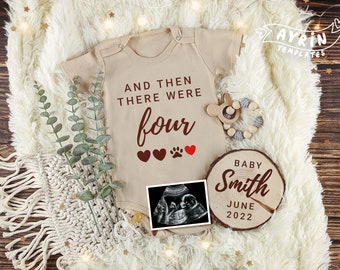 Könnte beinhalten: Ein beiger Strampler mit dem Text "And then there were four" und drei roten Herzen und einem Pfotenabdruck. Ein Holzschild mit dem Text "Baby Smith June 2022" und einem Schwarzweiß-Ultraschallbild.