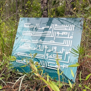 Op de afbeelding: Een tealblauw bord met witte Arabische kalligrafie erop. Het bord is gedeeltelijk bedekt met groen gebladerte en bruine dennennaalden.