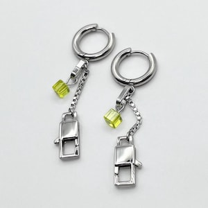 Op de afbeelding: Een paar zilverkleurige oorringen. Elke oorring heeft een kleine, vierkante, geelgroene kraal, een ketting en een rechthoekige bedel met een sluiting. De oorbellen worden tegen een witte achtergrond getoond.