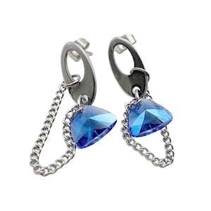 May include: A pair of silver-tone earrings featuring a triangular, faceted blue crystal pendant. Each earring has a chain that dangles from a silver oval shape. The earrings are set against a white background.