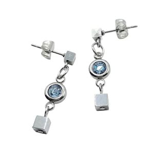 Può includere: Un paio di orecchini pendenti color argento. Ogni orecchino presenta un piccolo perno quadrato, una catena, una gemma blu chiaro incastonata in una lunetta rotonda, un'altra catena e un piccolo ciondolo quadrato. Su sfondo bianco.