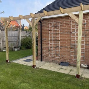 May include: A wooden pergola structure featuring vertical support posts anchored by dark metal bases, horizontal beams with notched ends, and diagonal corner braces. The structure is positioned against a red brick wall with two wooden lattice panels.