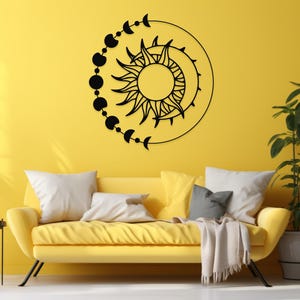 May include: Black metal wall art featuring a sun and moon design with phases of the moon. The artwork is circular and mounted on a yellow wall above a yellow sofa with white and gray pillows. Decorative home decor.