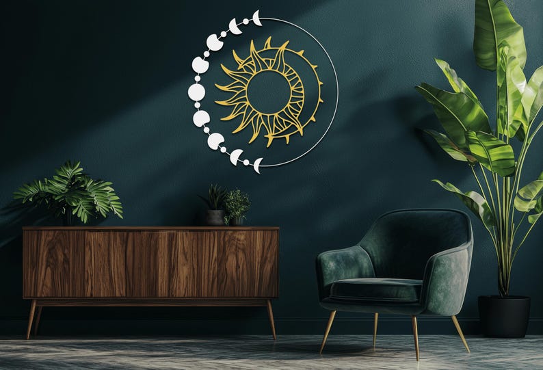May include: A celestial-themed wall art piece featuring a sun and moon design. The sun is gold, and the moon phases are white. The artwork is mounted on a dark teal wall, with a wooden cabinet, green chair, and plants in the foreground.