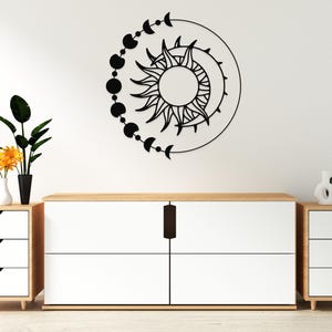 May include: Black metal wall art featuring a sun and moon design with phases of the moon. The artwork is mounted on a white wall above a white and wood-toned cabinet. Decorative vases and plants are also present.