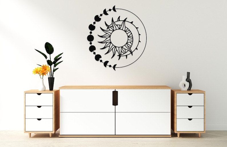 May include: Black metal wall art featuring a sun and moon design with phases of the moon. The artwork is mounted on a white wall above a white and wood-toned cabinet. Decorative vases and plants are also present.