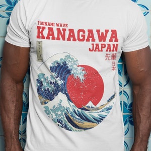 Puede incluir: Camiseta blanca con un gráfico rojo y azul de una gran ola y un sol rojo. El texto "TSUNAMI WAVE KANAGAWA JAPAN" está impreso en rojo en la camiseta. El texto japonés "先  日本" está impreso en negro en la camiseta.