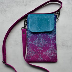 May include: A small, patterned crossbody bag with a teal flap and a magenta strap. The bag's body features a purple and blue geometric design. The strap is adjustable and attaches with silver-colored metal clips.