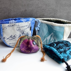 May include: Four fabric drawstring pouches of varying sizes and colors. One features a map with text reading Isengard and Minas, one has a blue mountain landscape, one is purple with gold script, and one is black and blue with a repeating pattern.