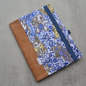 May include: A notebook cover with a brown leather-like spine and a blue and white patterned fabric cover. The fabric features cartoon characters and floral designs. A blue elastic band secures the notebook.
