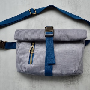 May include: A light gray corduroy fanny pack featuring a roll-top closure secured by a blue webbing strap and a plastic buckle. The bag includes a vertical front zipper pocket with a gold-toned metal carabiner and an adjustable blue webbing strap.
