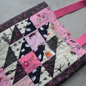 May include: A quilted tote bag with a Halloween theme. The bag features a patchwork design with pink, black, and white fabrics. The design includes images of ghosts, spiders, and haunted houses. The bag has pink straps and the text "Witch Please".
