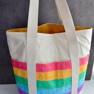 May include: A white canvas tote bag with rainbow colored stripes. The stripes are made of a patterned fabric with a topographic design. The bag has two long white canvas straps.