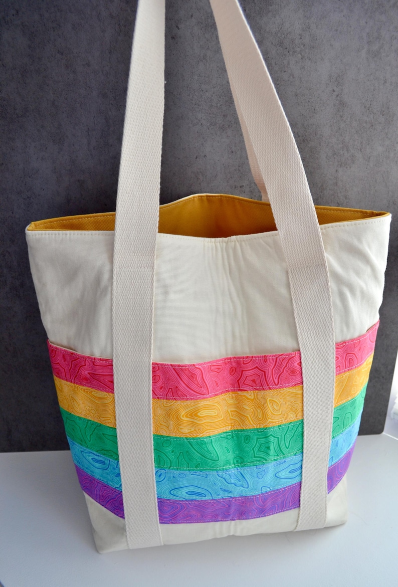 May include: A white canvas tote bag with rainbow colored stripes. The stripes are made of a patterned fabric with a topographic design. The bag has two long white canvas straps.