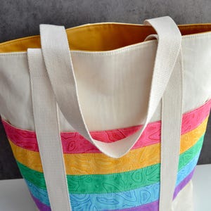 May include: A white canvas tote bag with rainbow striped fabric on the bottom. The bag has two long white straps and a yellow lining.