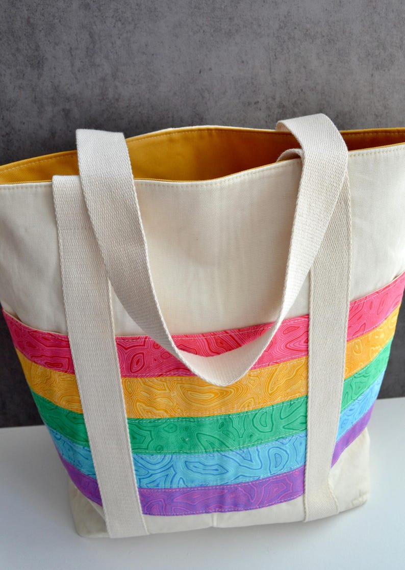 May include: A white canvas tote bag with rainbow striped fabric on the bottom. The bag has two long white straps and a yellow lining.