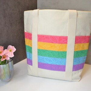 May include: A white canvas tote bag with rainbow colored stripes. The stripes are made of fabric with a floral pattern. The bag has two long straps.