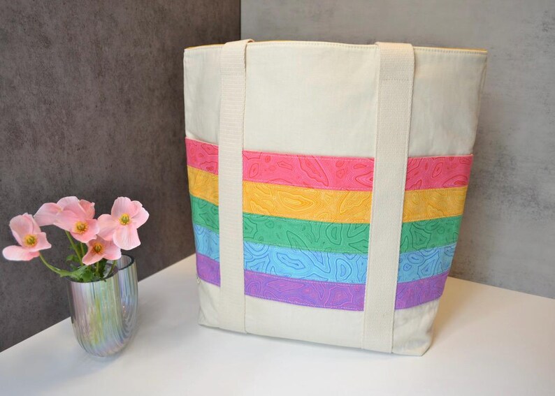 May include: A white canvas tote bag with rainbow colored stripes. The stripes are made of fabric with a floral pattern. The bag has two long straps.