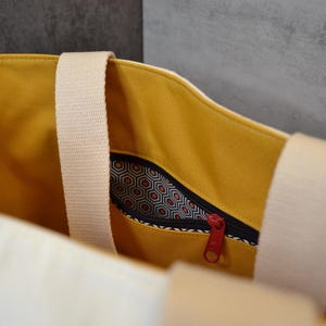 May include: A yellow canvas tote bag with a white zippered pocket and a white strap. The pocket has a black and white geometric pattern.