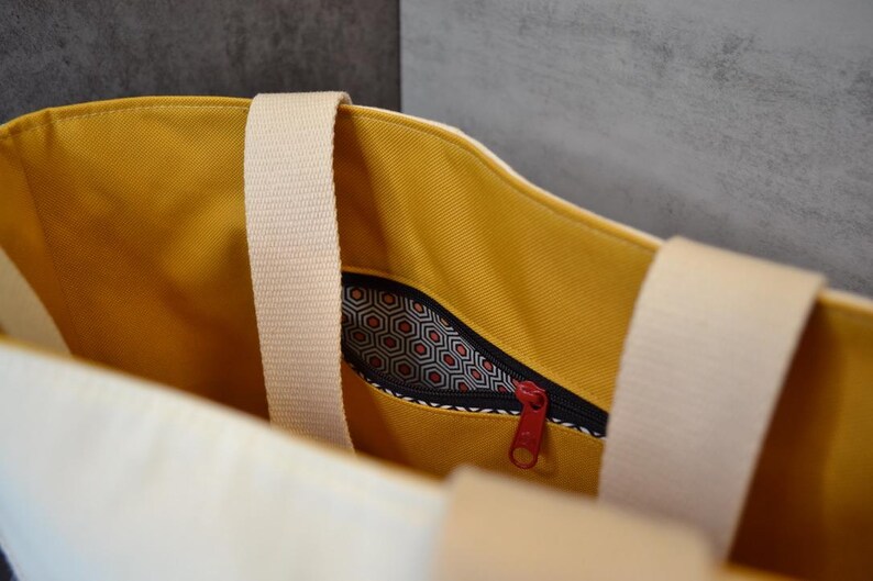 May include: A yellow canvas tote bag with a white zippered pocket and a white strap. The pocket has a black and white geometric pattern.