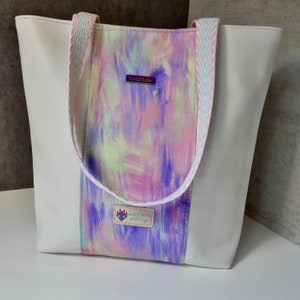 May include: White tote bag with a central panel featuring a colorful abstract design in shades of purple, pink, and yellow. The bag has white sides and a patterned strap. The text "silent bear stitching" is visible on a small label.