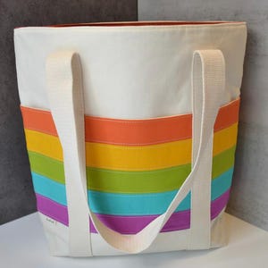 May include: White canvas tote bag with a rainbow stripe design. The bag features a rectangular shape with sturdy white handles. The rainbow stripes include orange, yellow, green, blue, and purple. The bag is designed for carrying items.