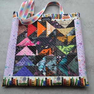May include: A handcrafted tote bag with a patchwork design featuring triangles in various colors, including red, orange, and purple. The bag has a light purple border and a colorful strap. The word "OCTOBER" is visible on the patchwork.