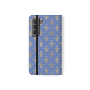 May include: A blue phone case featuring a repeating pattern of gold-colored bees and small crowns. The case has a vertical black elastic strap running along the left side and a rectangular camera cutout at the top left corner of the device.